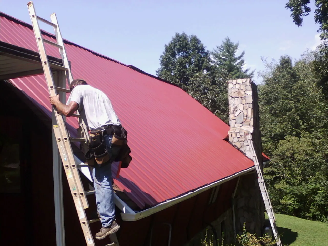 Roofer on a ladder beside a red metal roof and stone chimney — Soffit & Fascia Repair by Roofing Brothers in Fort Collins